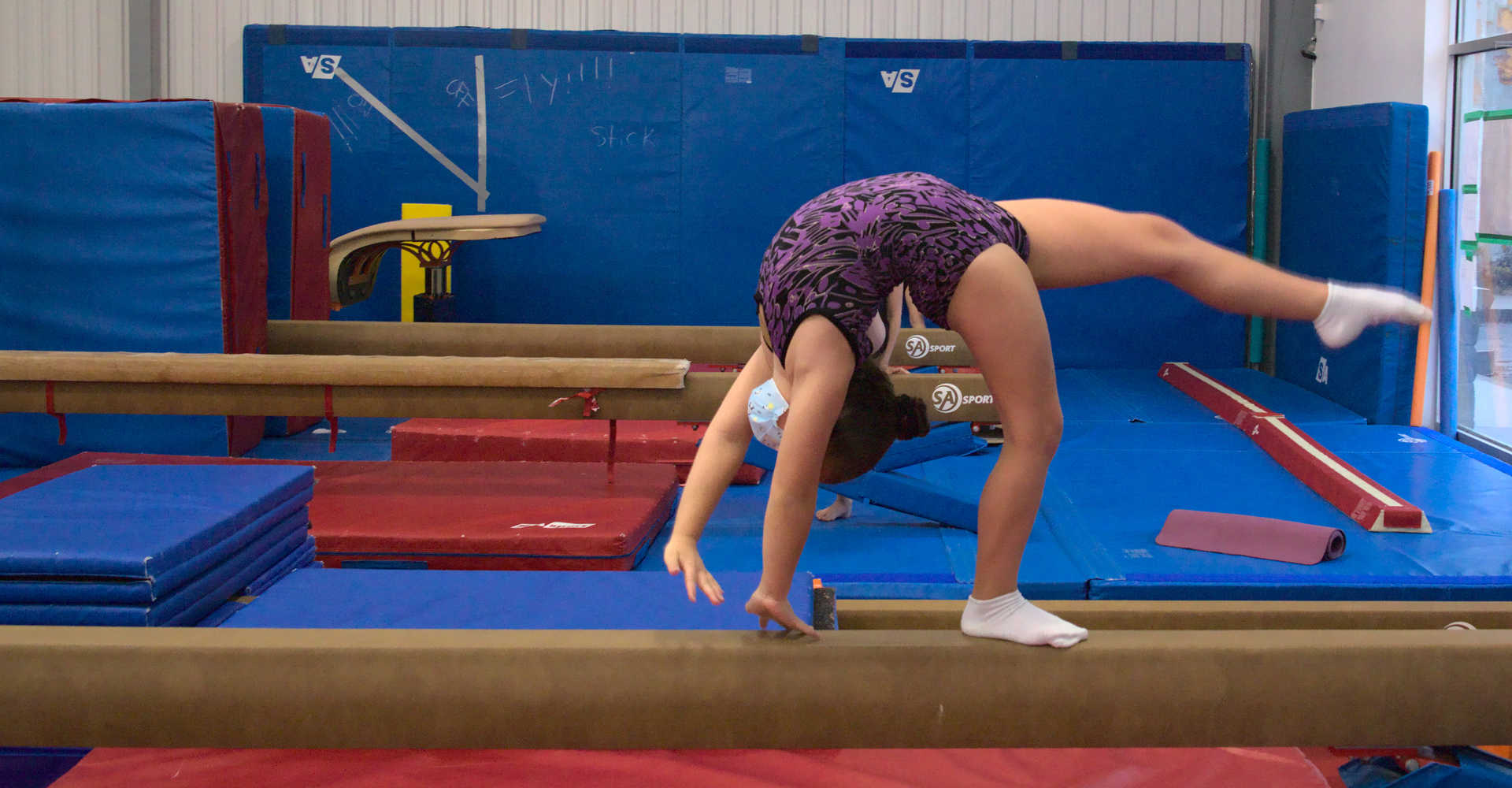 Young gymnast on a balance beam practicing a back walkover at Gymworld Adventures in Gymnastics facility in North London.