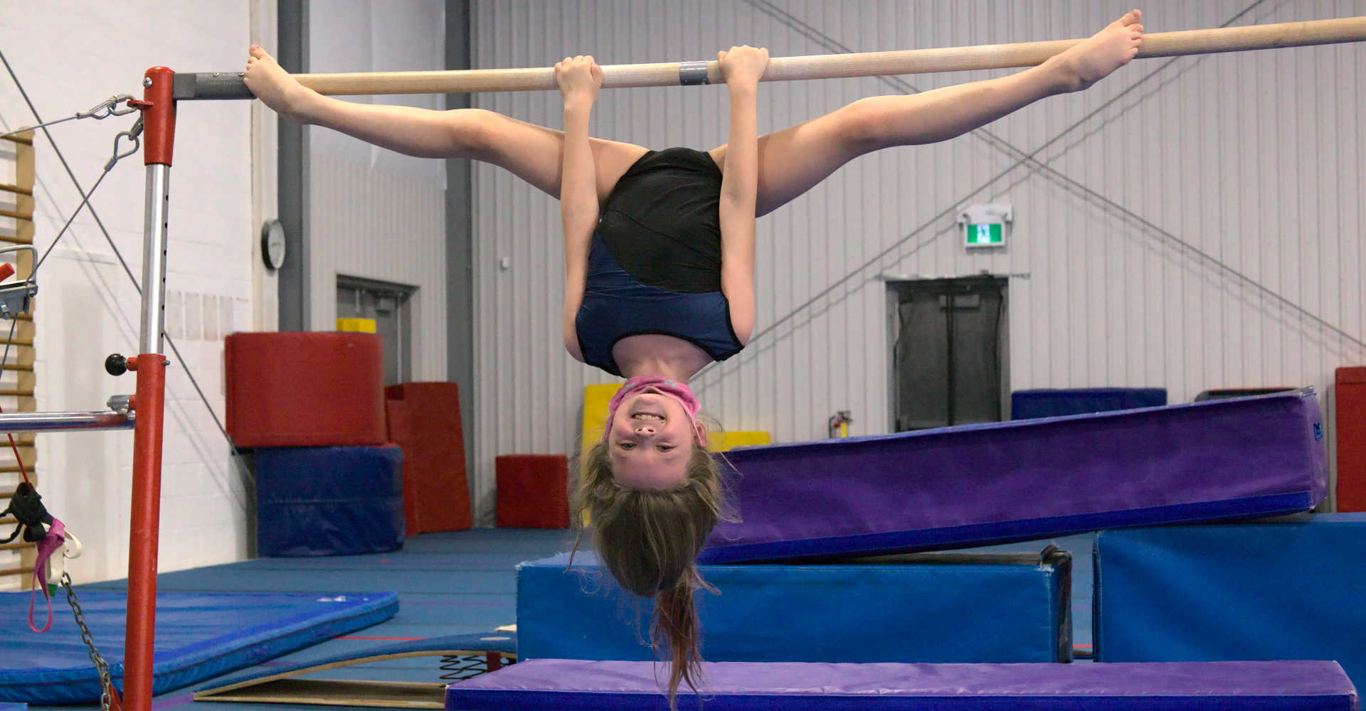 A smiling young gymnast, hanging upside down on the bar, takes a small break to catch a breath and strike a pose at Gymworld Adventures in Gymnastics in Northwest London